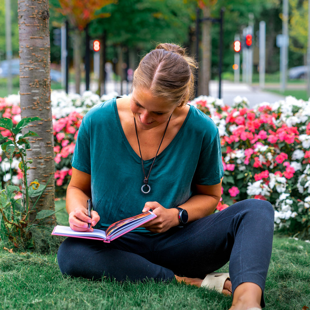Young woman writing in gratitude and manifestation journal while sitting on grass